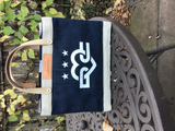 Unofficial DC × Apolis Petite Market Bag in navy jute with natural trim, tan leather handles and the white DC Love emblem with three stars on the front panel